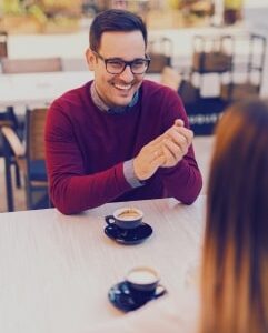 mężczyzna w swetrze w kolorze bordo siedzi przy stole i uśmiecha się do kobiety, na stole stoją dwie ciemne filiżanki kawy, w tle widoczne stoły restauracji, speed dating, Warszawa, spotkania do slingli w każdym wieku