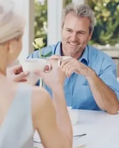 mężczyzna w błekitnej koszuli, siwe włosy siedzi w restauracji przy stole z blonynką w jasnoszarej bluzce, biały obrus, uśmiechnięci, randka spotkania dla singli, tinder, dreamdate, event dla Singli, szybkie randki dla dorosłych Warszawa