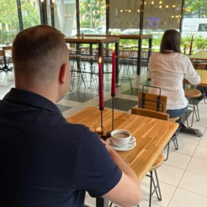 mężczyzna siedzi sam przy stole w kawiarni, drewniany stół. dalej widać kobietę siedzącą samotnie przy stole, randka speed dating, szybkie randki, Warszawa, mini randki, tinder, upoluj singla, randki na żywo, jak się przygotować na randkę, o czym rozmawiać na randce, szukanie miłości, obecne randki, randki na czasie, swatka, współczesna swatka ile czasu na randkę, czas na miłość