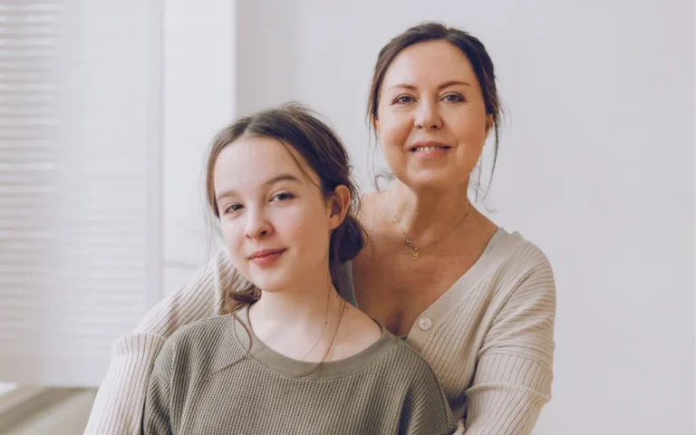 Uśmiechnięta kobieta obejmuje nastoletnią dziewczynę, obie patrzą w stronę obiektywu. Tło jasne i minimalistyczne, w domowym wnętrzu.