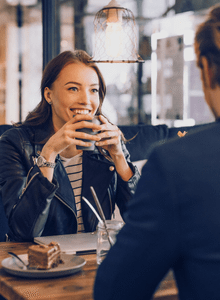 kobieta w czarnej skórzanej kurtce siedzi przy stole z mężczyzną, w kawiarni, kobieta trzyma w dłoni kubek, na stole stoi deser- ciasto na talerzy, na przeciwko siedzi mężczyzna, , nie widać twarzy, meżczyzna ubrany w ciemny swetr, szybkie randki u Amora , single Warszawa, co robić w Warszawie