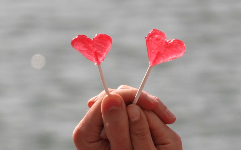 Dwie splecione dłonie kobieca i męska trzymajce po jednym lizaku na patyku w kształcie czewonego serca niewielkich rozmiarów. W tle zamazany obraz wody z przebijającym motywem małych fal. Całość symbolizuje walentynki i miłość