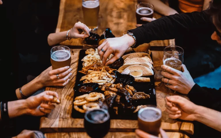 Stół drewniany w klimatycznej restauracji, wokół którego siedzi kilka osób - kobiet i mężczyzn, ale wydać tylko ich ręce. Na stole stoi duża czarna taca z przekąskami jak frytki, taco i burgery oraz szklanki z piwem. Osoby wokół stołu częstują się.